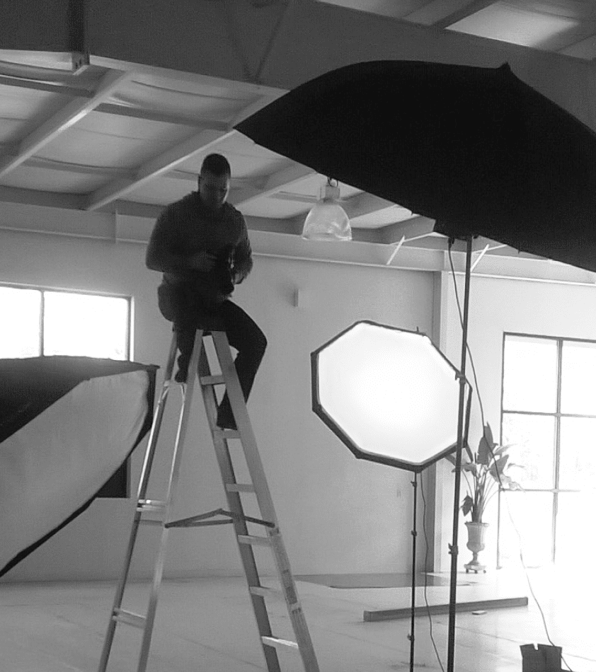 A picture of Andrew Sears in a studio sitting on a ladder, looking at his camera